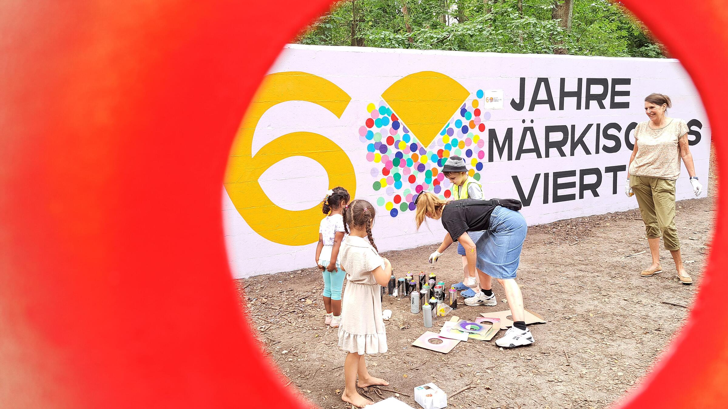 Außenaufnahme zeigt vier Personen, darunter zwei Kinder, beim Besprühen der Graffitiwand, durch eine runde Schablone fotografiert.