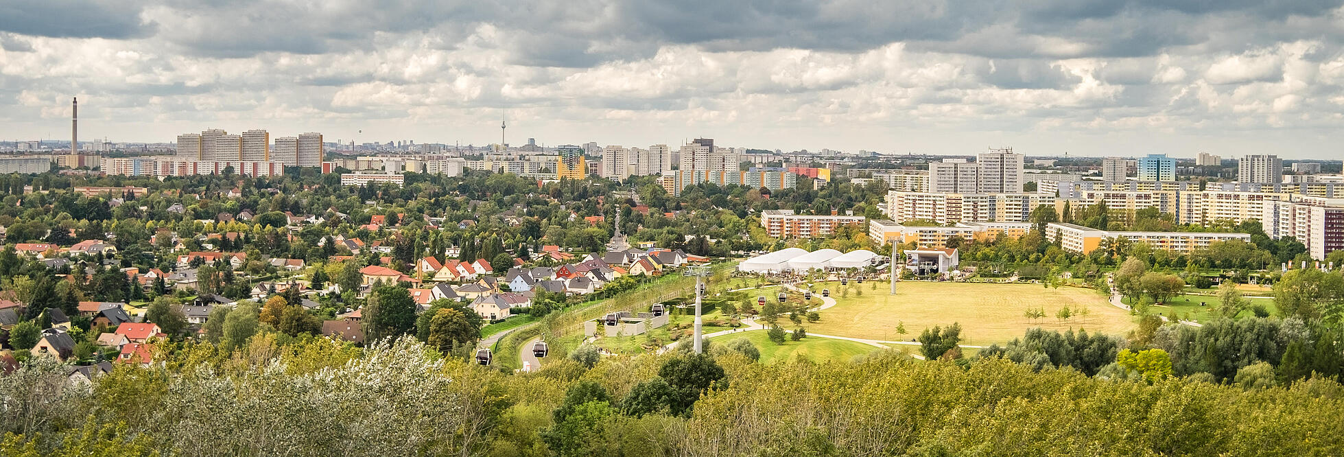 Die Außenaufnahme zeigt eine Panoramaaussicht über den Berliner Bezirk Marzahn-Hellersdorf vom Kienberg aus. Über den zahlreichen Wohnhäusern sammeln sich Wolken am Himmel. Zwischen den Häuserreihen befinden sich viele grüne Bäume. Ganz in der Ferne kann man das berühmte Berliner Wahrzeichen, den Fernsehturm, erspähen.