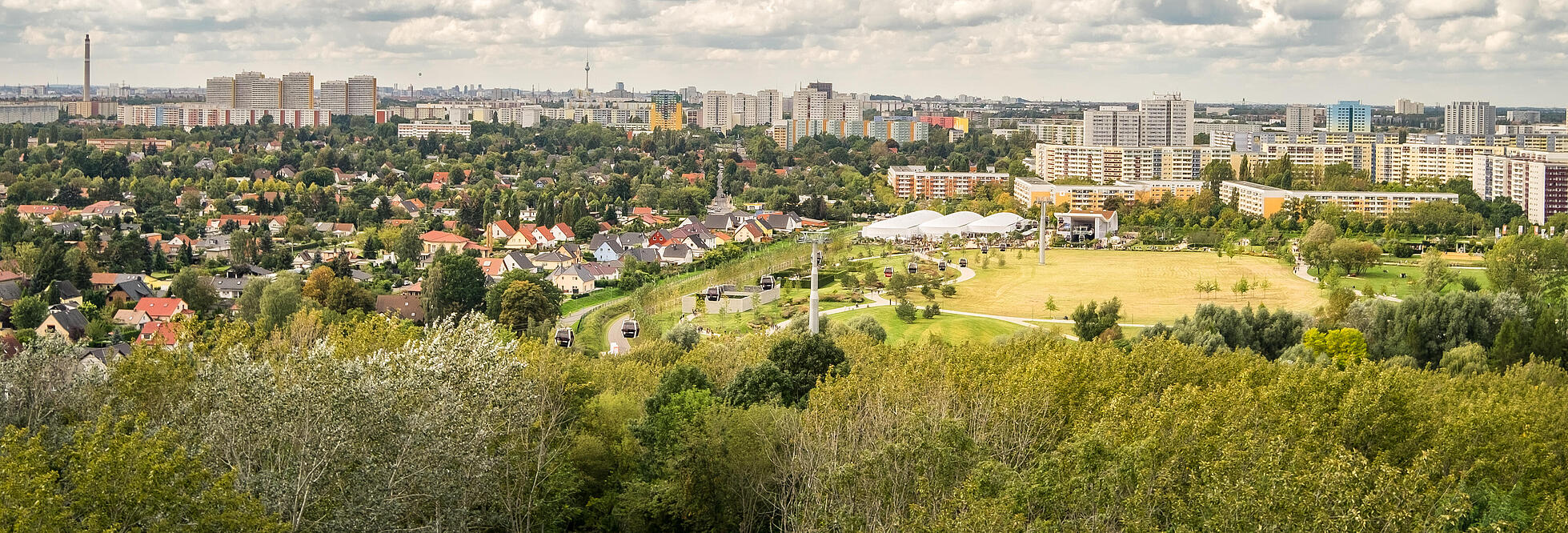 Die Außenaufnahme zeigt eine Panoramaaussicht über den Berliner Bezirk Marzahn-Hellersdorf vom Kienberg aus. Über den zahlreichen Wohnhäusern sammeln sich Wolken am Himmel. Zwischen den Häuserreihen befinden sich viele grüne Bäume. Ganz in der Ferne kann man das berühmte Berliner Wahrzeichen, den Fernsehturm, erspähen.