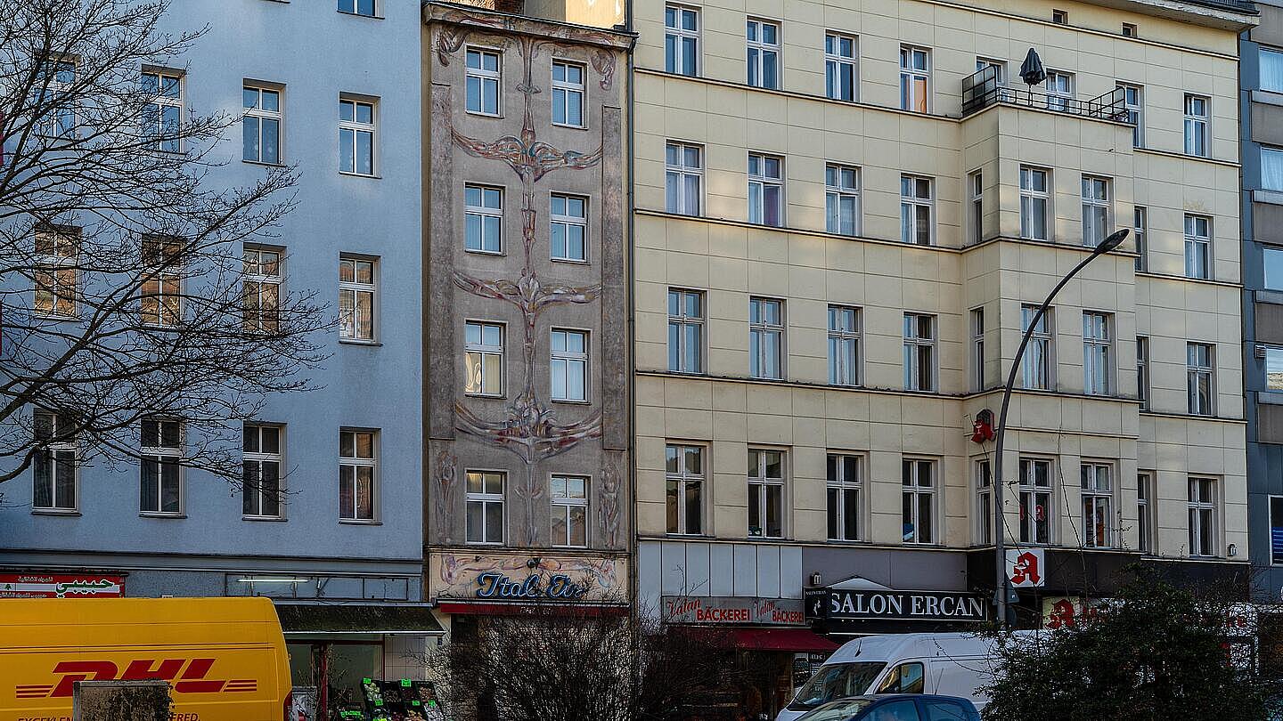 Die Außenaufnahme zeigt die Fassade einer Häuserreihe in Berlin-Wedding in der Müllerstraße. Eine blaue, graue und gelbe Fassade reihen sich aneinander. Im Erdgeschoss der Häuser befinden sich ein Bäcker, ein Restaurant und eine Apotheke. Vor den Häusern parken Autos. Auf der linken Seite lässt sich ein DHL-Lieferfahrzeug erkennen.