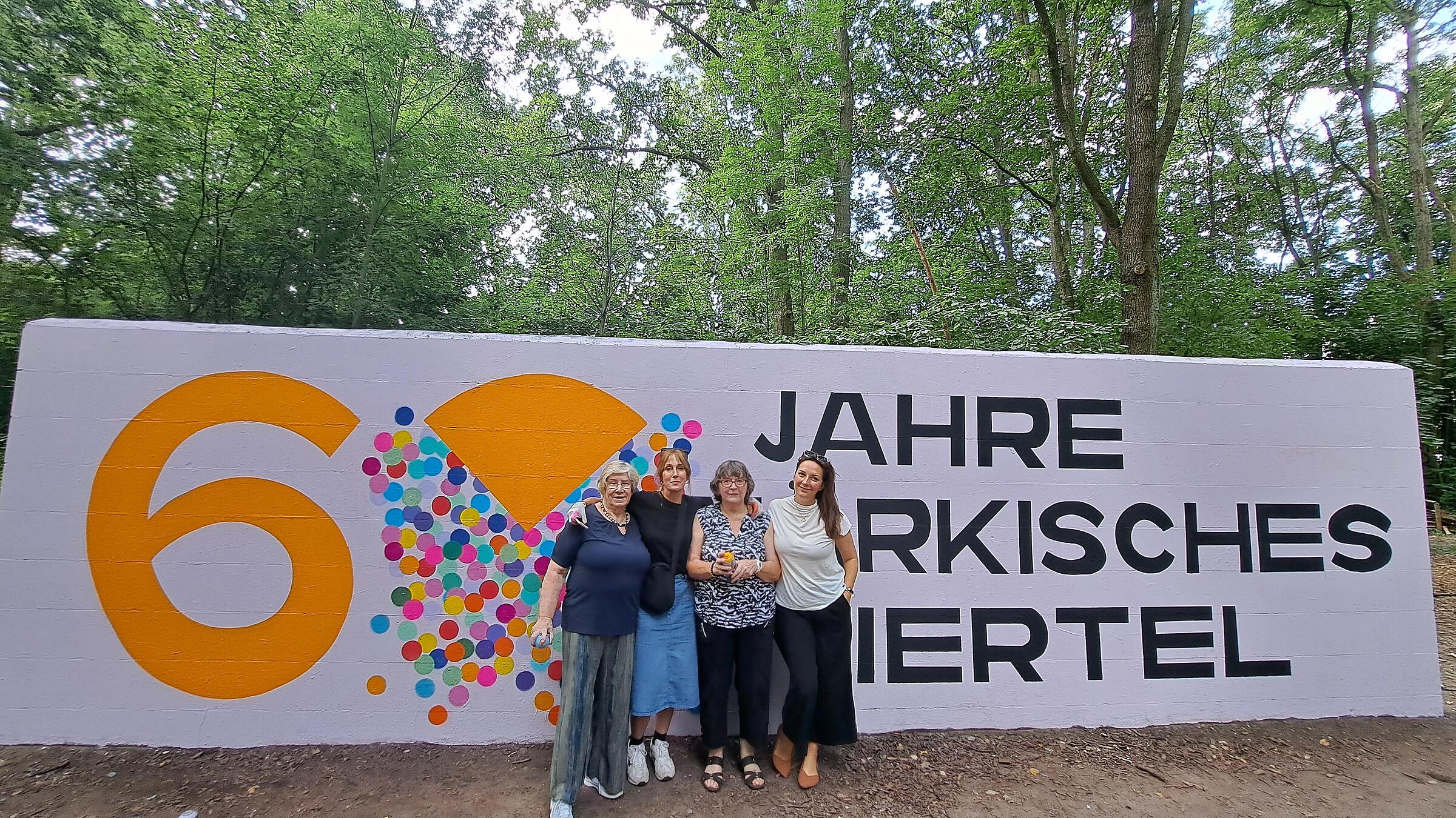 Außenaufnahme zeigt vier Frauen unterschiedlichen Alters, die lächelnd vor der Graffitiwand mit dem Logo des Märkischen Viertel stehen.