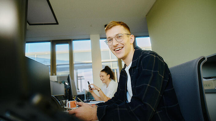 Innenaufnahme zeigt zwei Mitarbeitende der GESOBAU bei der Arbeit. Der Kollege im Vordergrund sitz an einem Schreibtisch und lächelt in die Kamera.