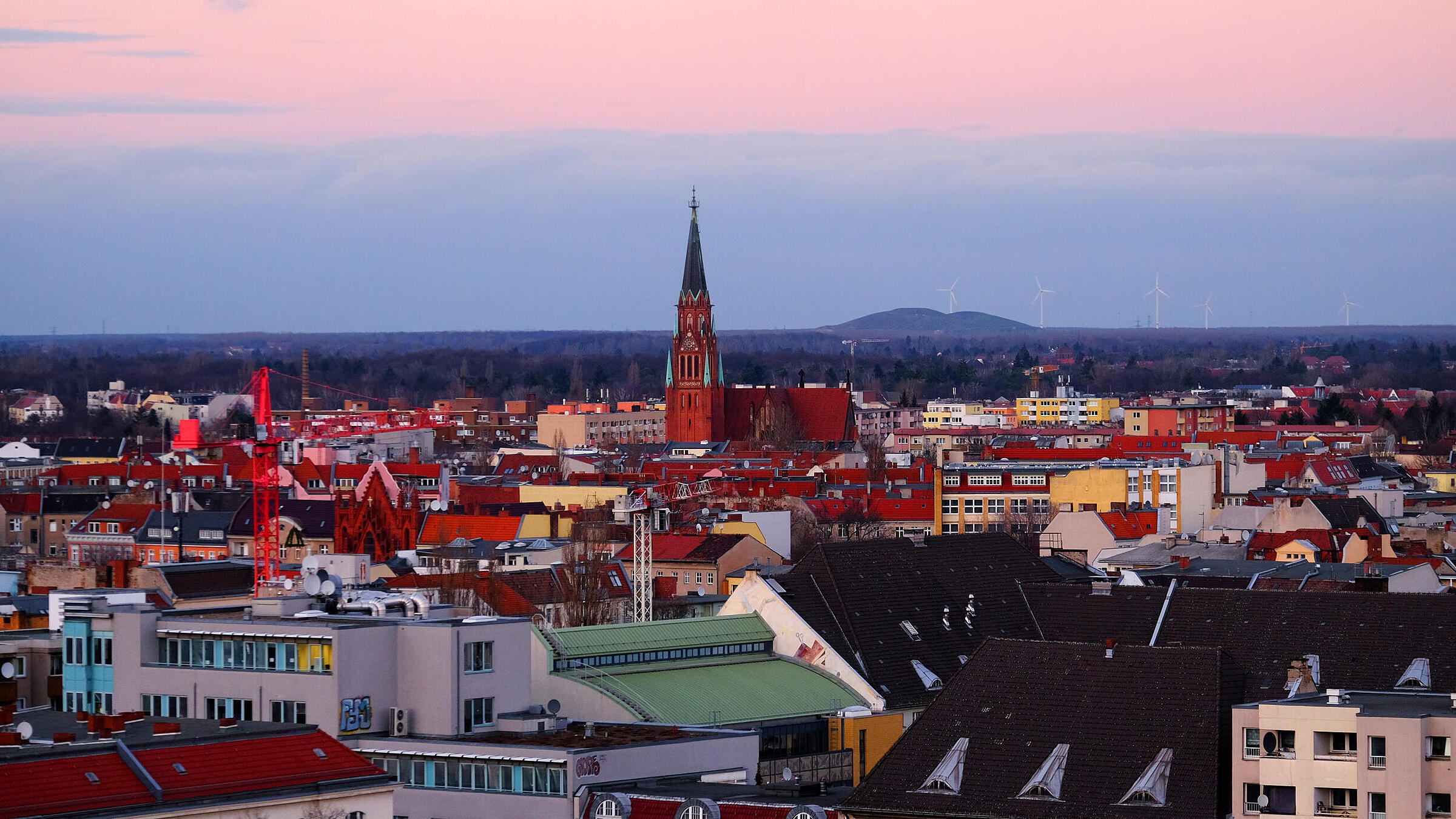 Die Außenaufnahme zeigt den Ausblick auf den Bunkerberg von Berlin-Wedding bei Sonnenuntergang. Die Farbpalette der Aufnahme erstreckt sich in warmen Rot- und Rosetönen. Im Hintergrund lassen sich in weiter Ferne Windräder erkennen. Der Großteil des Bildes zeigt jedoch das Dächermeer von Berlin-Wedding.