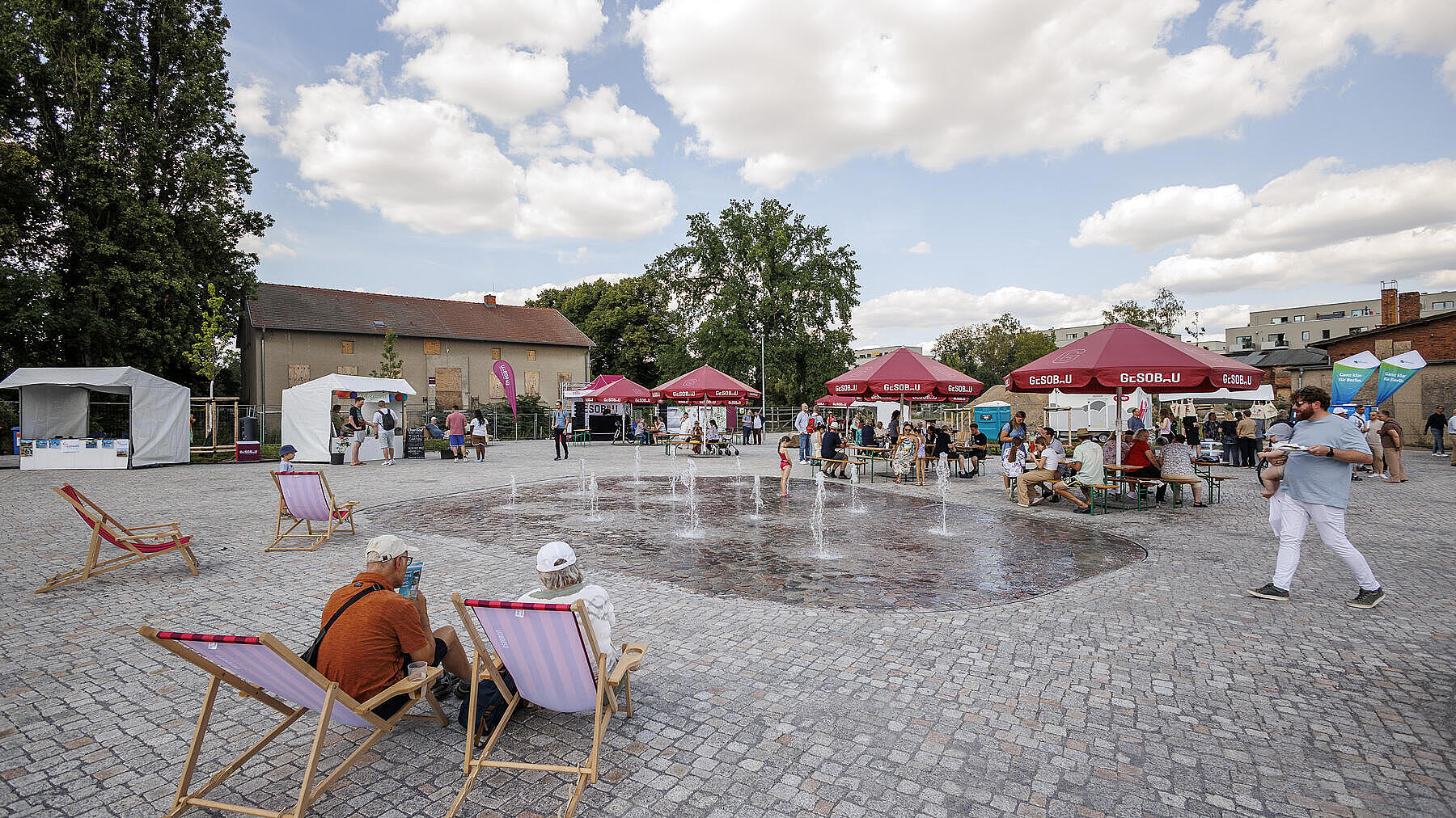 Die Außenaufnahme zeigt den Stadtplatz in Berlin Hellersdorf. In der Mitte des Platzes befinde sich ein Brunnen mit mehreren kleinen Fontänen. Um den Platz herum befinden sich Personen auf Bierbänken oder Liegestühlen. Im Hintergrund zu erkennen sind vereinzelte Markt- und Informationsstände.