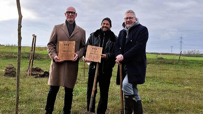 Bild zeigt  Jörg Franzen (Vorstandsvorsitzender GESOBAU AG), Jan Kinappel (Geschäftsführer Anschütz Entertainment Group Deutschland (AEG)), Jan Schierhorn (Initiator FAME FOREST) bei Kickoff für den FAME Forest Berlin. im Hintergrund Acker, im Vordergrund geplanzter Baum. Protagonisten halten Ehrentafel in der Hand.