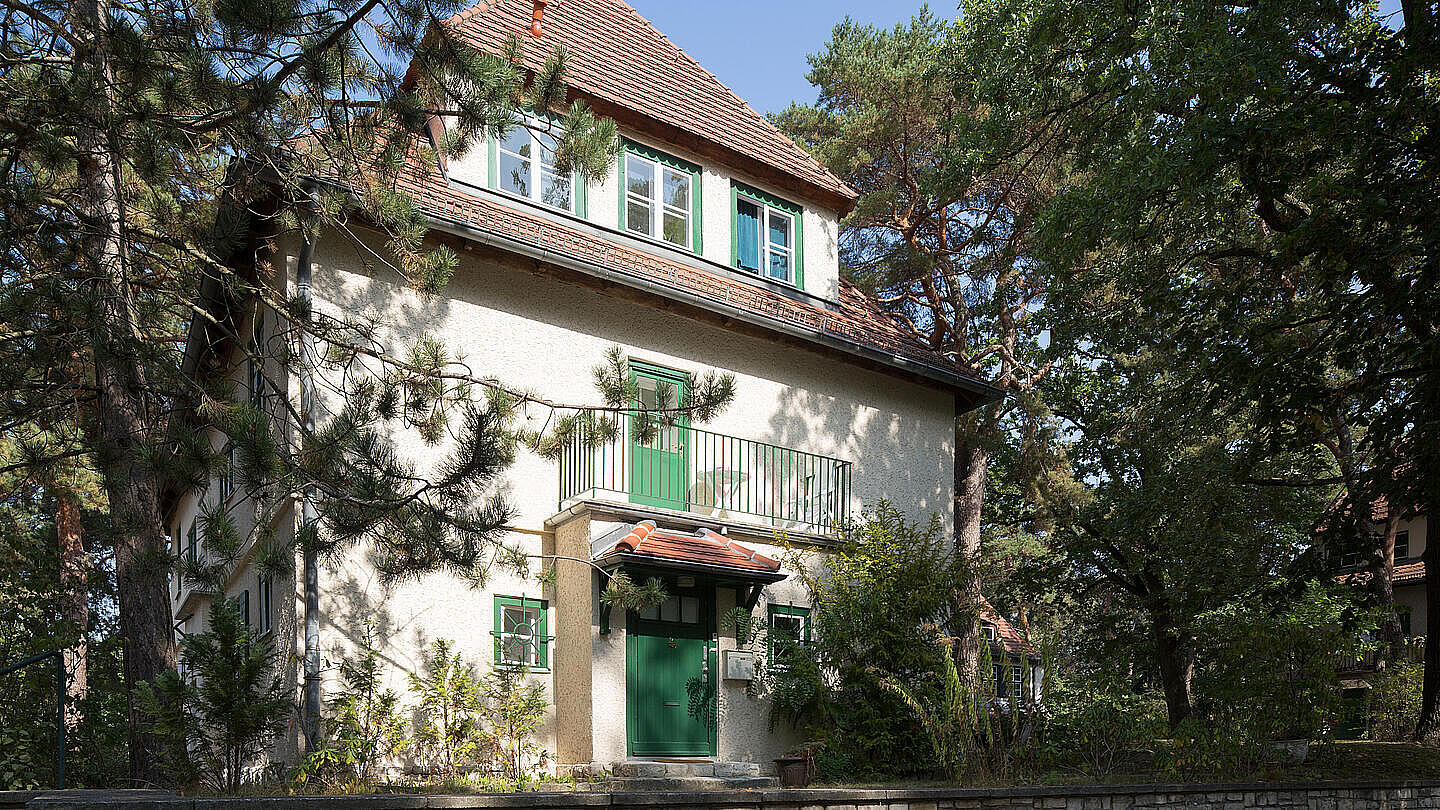 Die Außenaufnahme zeigt eine Bestandswohnung der GESOBAU in der Reinerzstraße. Um das Haus herum befinden sich viele Bäume und Sträucher. Die Sonne scheint. Das Haus verfügt über einen Balkon.