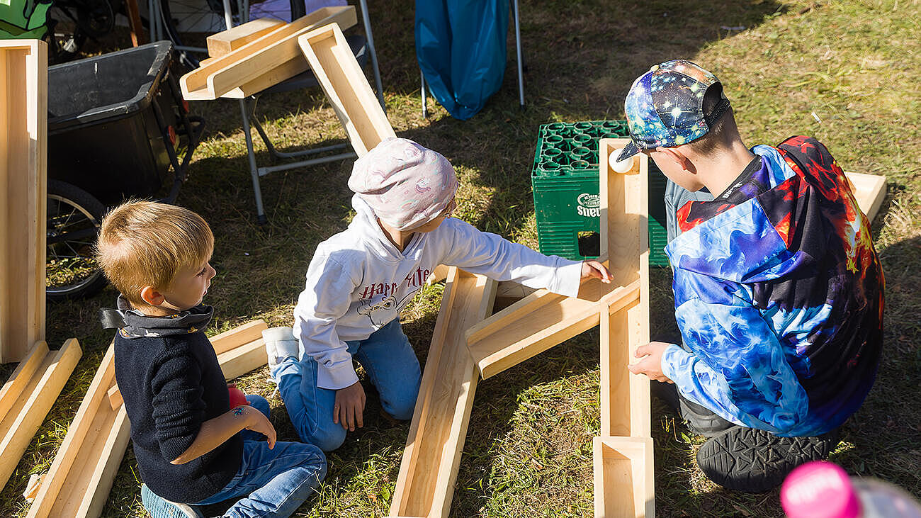 Außenaufnahme zeigt drei im Gras sitzende Kinder, die eine Riesenmurmelbahn aus Holzteilen zusammenbauen.