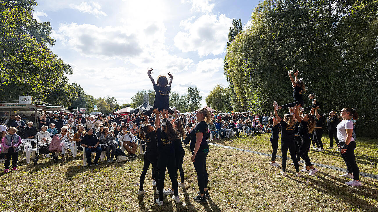 Gruppe sehr junger Cheerleaderinnen bei einer Hebefigur, im Hintergrund die Zuschauermenge im Sonnenschein. 