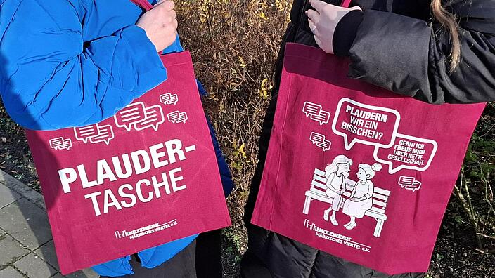 Bild zeigt zwei Personen, sich zugewandt mit jeweils einer Plaudertasche (Jutebeutel mit Print).