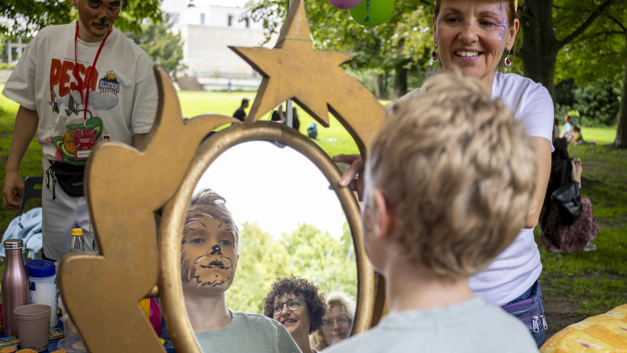 Ein im Gesicht als Tiger geschminktes Kind betrachtet sich unter den Augen von Erwachsenen in einem Spiegel. Wir sehen sein Gesicht im Spiegelbild und die zwei Erwachsenen, die für das Schminken verantwortlich sind, links im Bild ein als Raubkatze geschminkter Mann, rechts eine Frau mit Glitzerornamenten an ihren Augen.