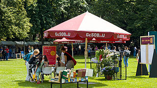 Außenaufnahme zeigt einen großen Sonnenschirm auf der Festwiese, darum gruppiert Bänke und Metaplanwände.