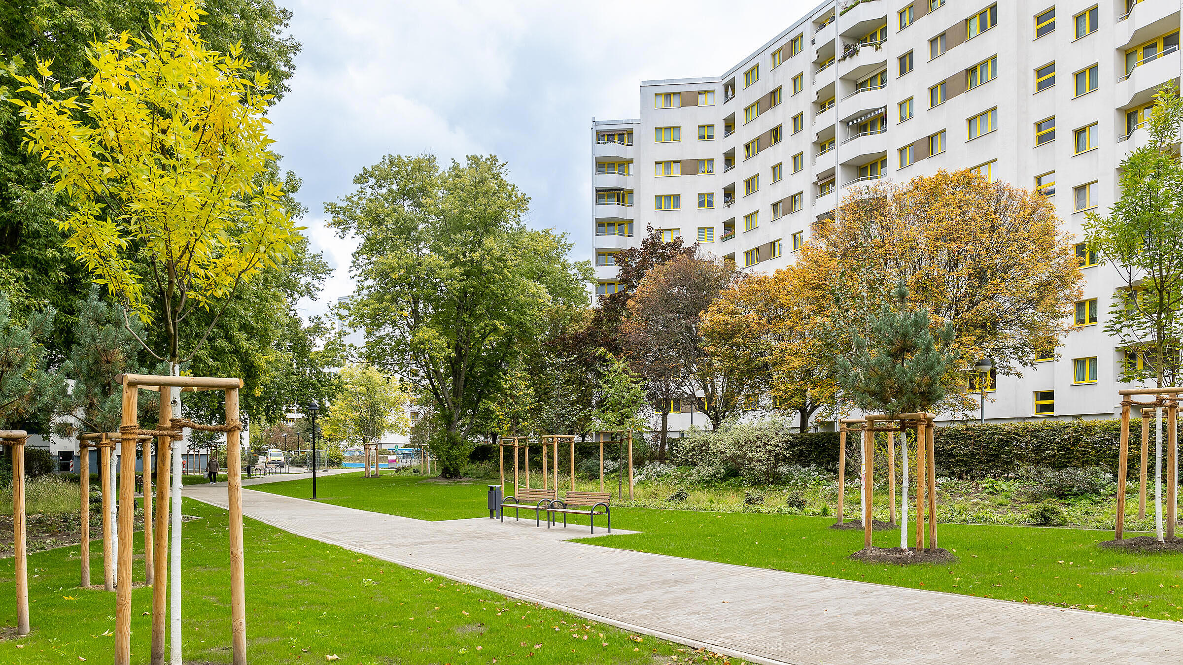 Die Außenaufnahme zeigt die Außenanlagen des Märkischen Viertels in Berlin Reinickendorf. Vor den sich im Hintergrund befindlichen weißen Fassaden ist ein Weg umringt von grüner Rasenfläche zu sehen. Zusätzlich zieren Bäume und Sitzgelegenheiten die Rasenfläche.