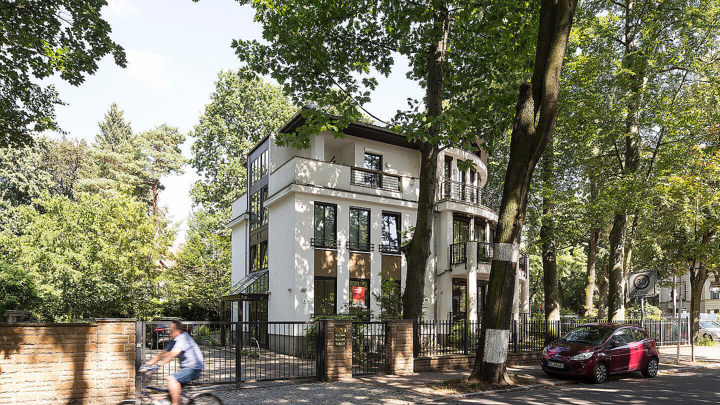 Die Außenaufnahme zeigt einen Bestand der GESOBAU in der Douglasstraße. Das alleinstehende moderne Haus mit weißer Fassade ist umringt von viel Grün. Auf der Straße davor parkt ein rotes Auto. Ein Fahrradfahrer fährt vorbei.