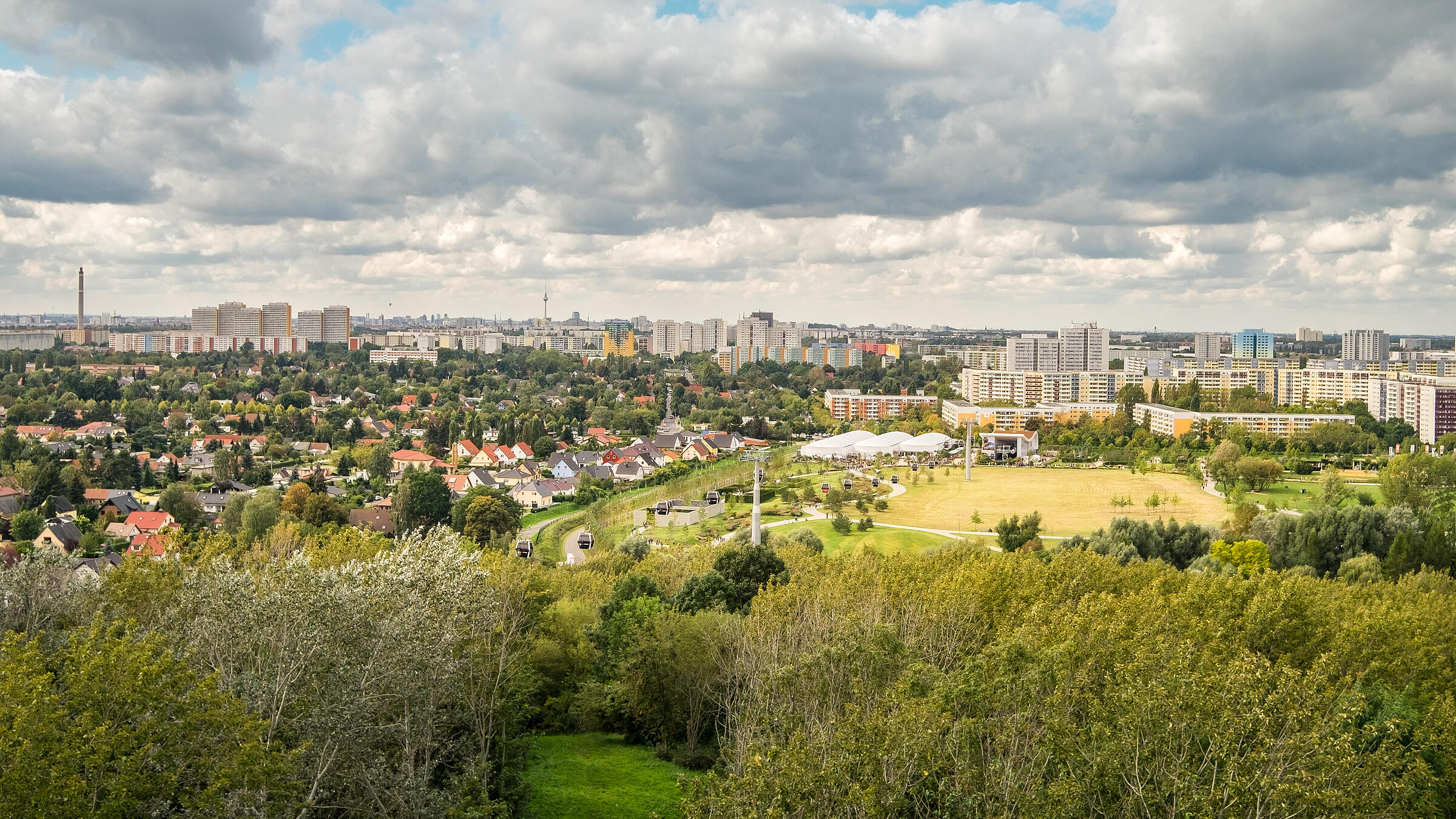 Die Außenaufnahme zeigt eine Panoramaaussicht über den Berliner Bezirk Marzahn-Hellersdorf vom Kienberg aus. Über den zahlreichen Wohnhäusern sammeln sich Wolken am Himmel. Zwischen den Häuserreihen befinden sich viele grüne Bäume. Ganz in der Ferne kann man das berühmte Berliner Wahrzeichen, den Fernsehturm, erspähen.