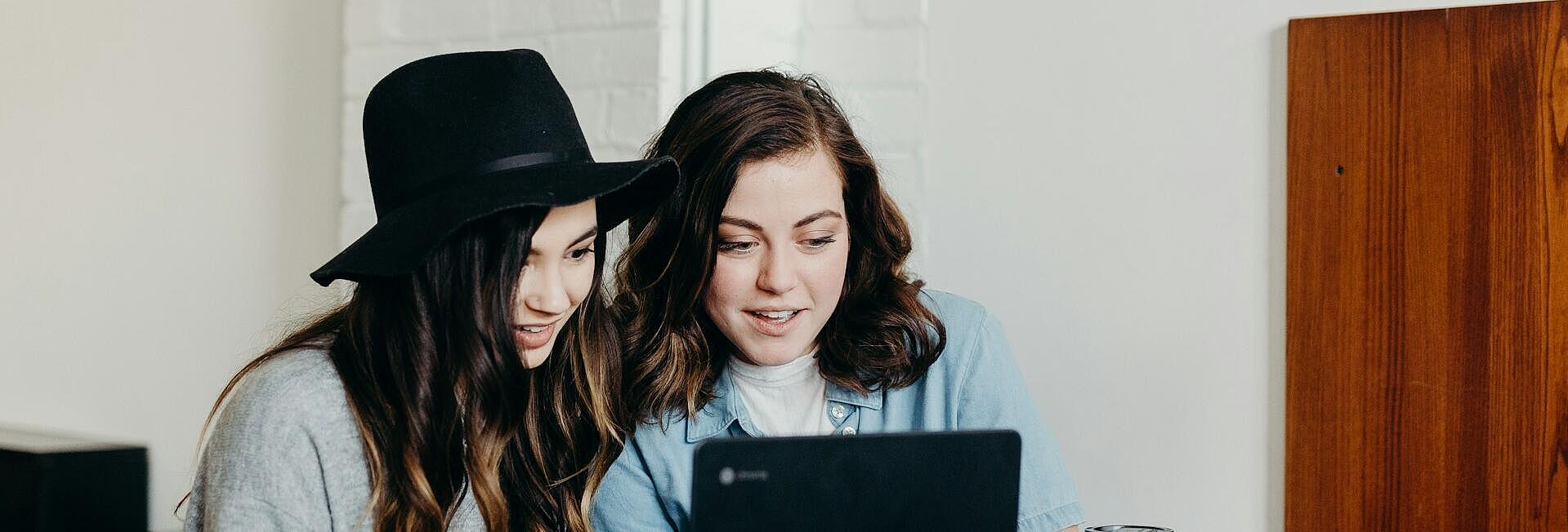 Zwei junge Frauen sitzen vor einem augeklappten Laptop an einem Holztisch.