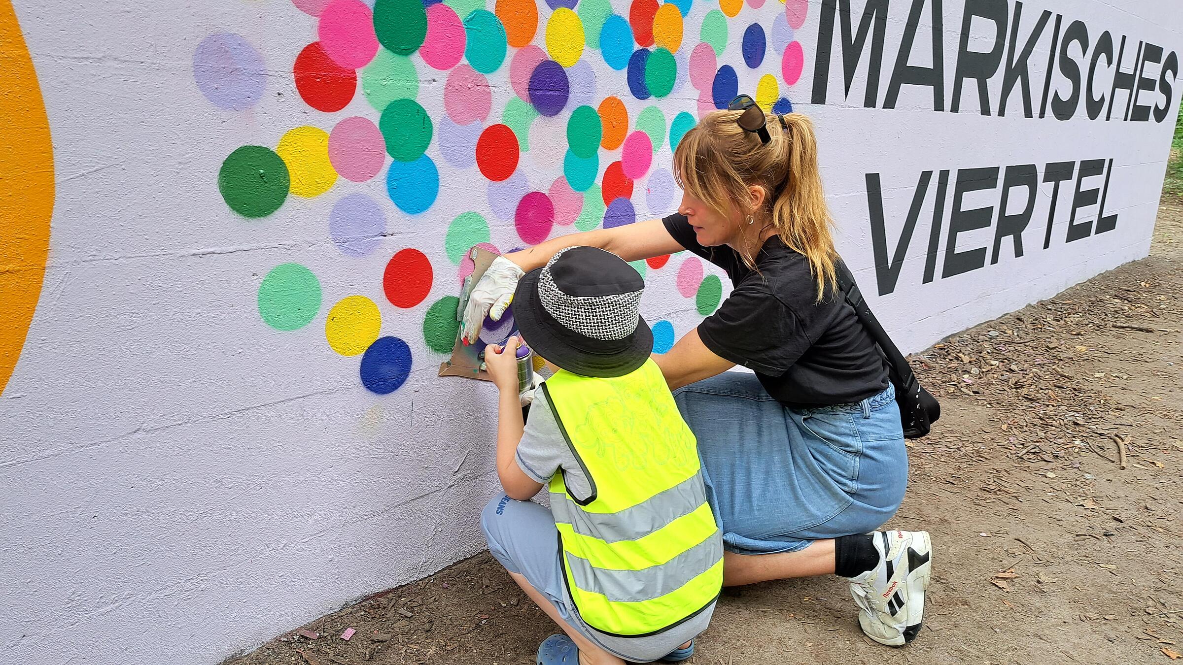 Außenaufnahme zeigen ein Kind und eine Erwachsene in der Hocke beim Sprayen von Farbklecksen als Teil des Jubiläumslogos.