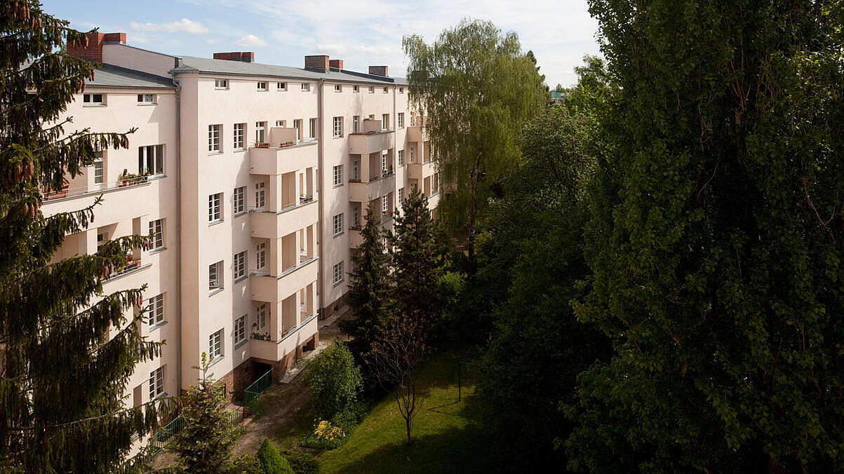 Die Außenaufnahme zeigt die rosaferbene Fassade mehrer Mehrfamilienhäuser aus der Vogelperspektive. Im Vordergrund der Häuser befindet sich grüner Rasen sowie etliche Sträucher und kleine sowie große Bäume. 