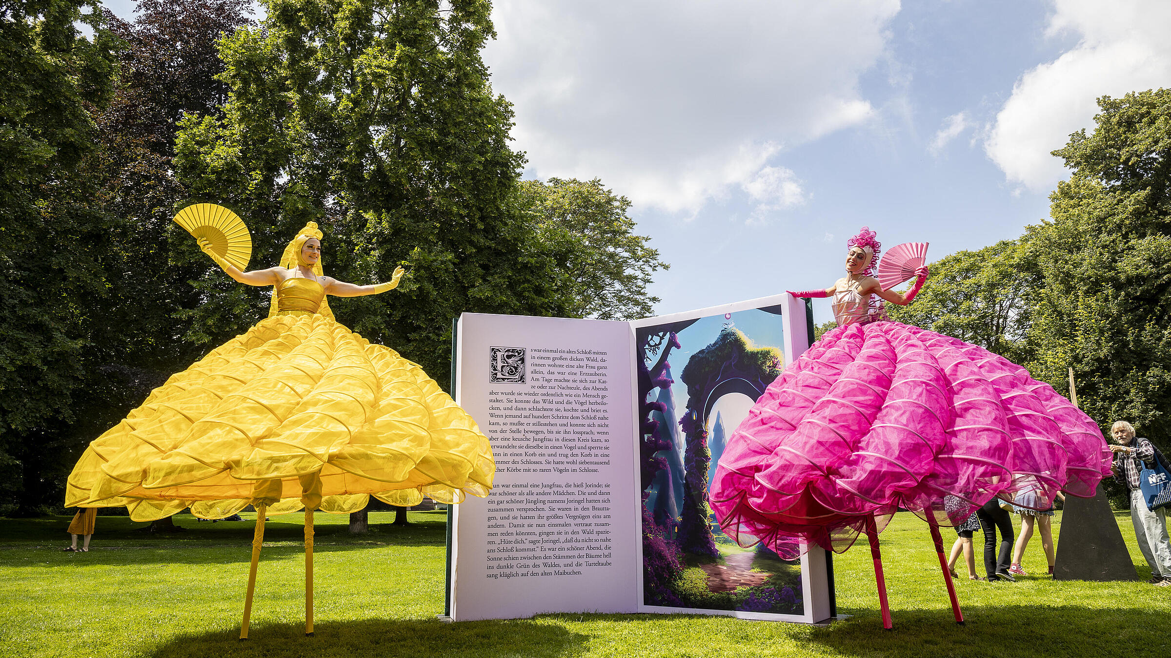 Aufnahme zeigt zwei Stelzenläuferinnen in voluminösen Kleidern und mit Fächern auf der Wiese vor dem Schloss. Beide flankieren gigantisches, aufgeklapptes Märchenbuch, das als Skulptur für das Kunstfest aufgestellt wurde.