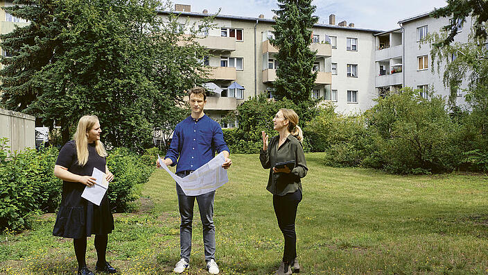 Außenaufnahme zeigt eine Gruppe von drei Mitarbeitenden der GESOBAU in einem grünen Innenhof stehend. Die Person in der Mitte hält einen aufgefalteten Plan in seinen Händen, seine Kolleginnen, ihm zur Seite stehend, sind im Gespräch. 