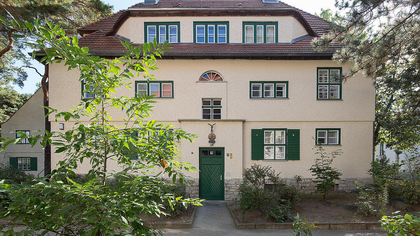 Die Außenaufnahme zeigt eine Bestandswohnung der GESOBAU mit grüner Haustür und grünen Fensterrahmen in der Reinerzstraße. Des Gebäude ist umgeben von grünen Sträuchern und Bäumen.